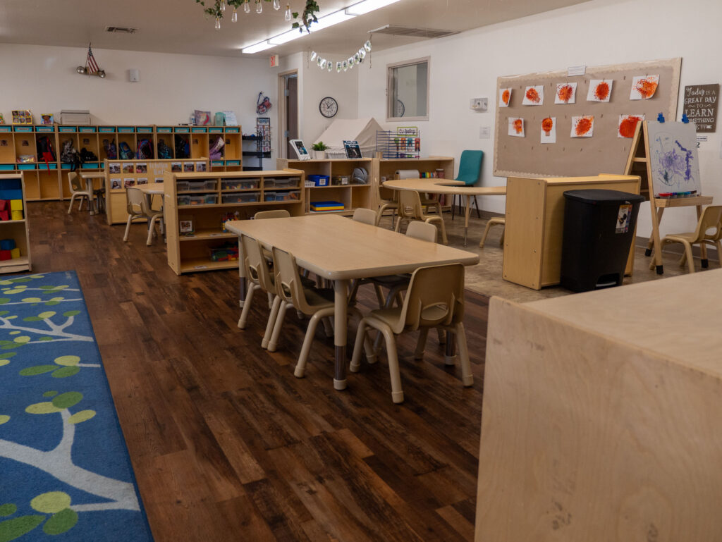 preschool classroom with tables, art display wall, and center shelves