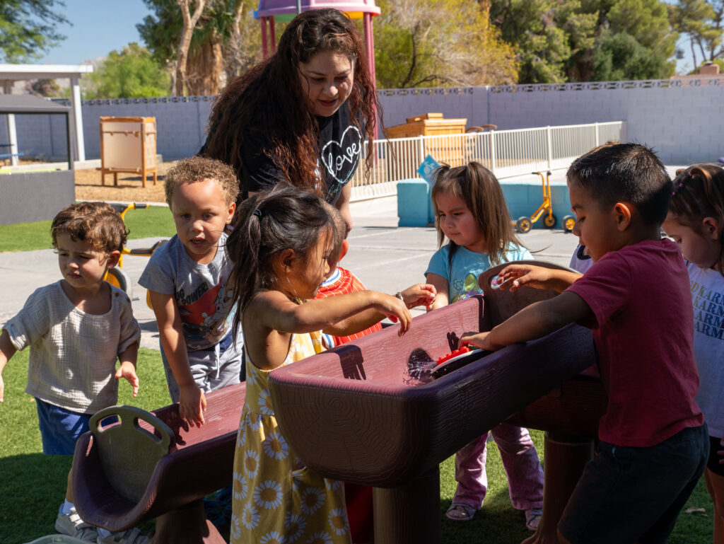 team \ our facility\ preschool teacher guiding children at outdoor water play table